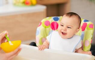 A mom is feeding her baby boy in a high chair in the kitchen. The mood is warm and affectionate, with a cozy kitchen environment enhancing the loving scene. photo