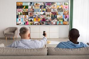 Two men are sitting on a couch in a living room, watching a large TV screen displaying a grid of various images. The man in the foreground is holding a remote control and is looking at the TV. photo