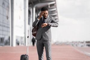 An African American businessman with luggage is checking the time outside a modern airport. The scene is professional and busy, with the sleek architecture of the airport in the background. photo