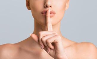 A closeup photo of a woman face with her finger pressed to her lips. She is making a shhh gesture, which indicates that she wants to be quiet. The background is a plain grey, cropped