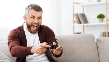Positive man is seated on a couch, holding a game controller in his hands. He appears focused on the screen, engaged in a gaming session. photo