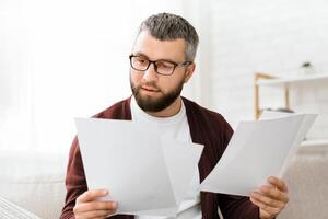 A man is seated on a couch, holding a stack of papers in his hands. He appears focused on the documents, possibly reading or reviewing them. photo
