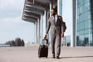 African American businessman is walking with his luggage outside a modern airport. The mood is dynamic and professional, highlighting travel and business with a sleek airport backdrop. photo
