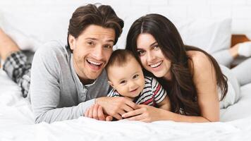 This image shows a happy family of three, a man, a woman, and a baby, lying in bed together. The baby is in the middle, with the parents on either side of him. photo