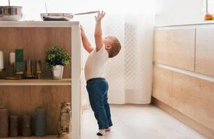 A curious toddler stands on tiptoe in a modern kitchen, reaching for a frying pan on the counter, illuminated by morning sunlight. photo