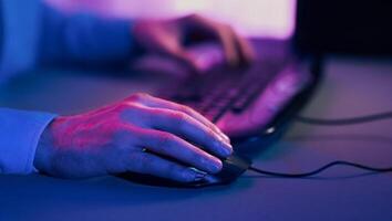 A persons hand is shown resting on a computer mouse, which is connected to a computer. The room is lit with blue and purple hues, creating a dramatic and colorful effect photo