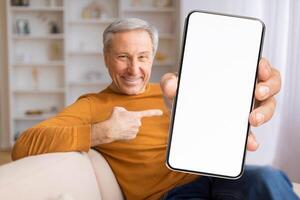 A cheerful senior man sits comfortably on a couch, pointing at a smartphone in his hand. The cozy living room offers a warm and inviting backdrop, emphasizing connection and technology. photo