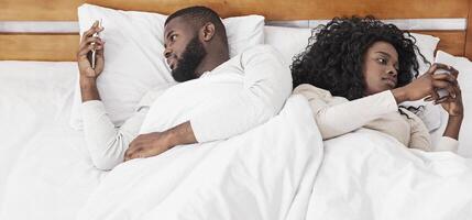Millennial couple ignoring each other after argue, lying in bed back to back and using their smartphones photo