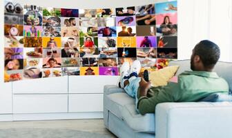 A man relaxing on white couch, his feet up, holding a remote control and watching a television screen. The television is displaying a unique grid of many small screens, each showing a different image photo