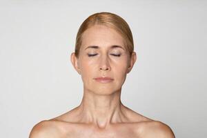 This is a portrait of a woman with blonde hair, wearing no makeup. She has her eyes closed and is looking straight at the camera. Her bare shoulders are visible, and her skin is fair photo