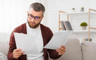 A professional man with glasses and a beard carefully reviews papers in a well-lit, modern home office setting during the day. He is seated on a sofa, concentrating on the documents in his hands photo