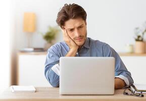 A man sits at a desk with a laptop in front of him, looking concerned. He rests his head on his hand while looking at the screen. photo
