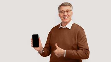 Mobile App. Smiling Elderly Man Showing Phone With Blank Screen Gesturing Thumbs Up Standing Over Gray Studio Background. Panorama, Mockup photo