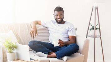 Black man in a white t-shirt and jeans sits comfortably on a couch in a living room. He is holding a white mug and smiling at the camera. There is a laptop on the coffee table in front of him photo