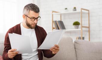 A man is seated on a couch, attentively reading a piece of paper in his hands. His concentration is evident as he scans through the document, absorbed in its contents. photo