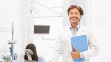 Visit my clinic. Handsome dentist keeping smile on his face and holding folder over cabinet background, empty space photo