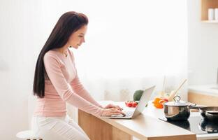 A woman is seated at a kitchen counter, engaged in using a laptop computer. She is focused on the screen, with her hands typing on the keyboard. The kitchen environment is visible in the background. photo