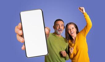 This image features mature couple posing excitedly while holding a smartphone with a blank screen in front of a purple background photo