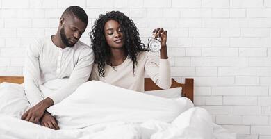 We hate mornings. Black married couple having problems with waking up, sitting sleepy in bed with alarm clock in hands, panorama with copy space photo