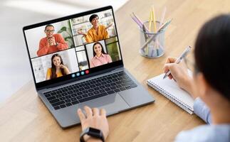 Woman takes notes on a notepad while participating in a conference meeting on a laptop computer. The screen displays four people in separate frames photo