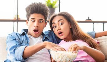 Black teen couple sits together on a couch, eyes wide with excitement as they watch an exciting movie. They are both holding popcorn bowls and enjoying the film photo