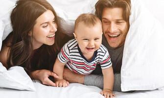 A happy family is playing peekaboo under a white blanket in bed. The parents are smiling and looking at their baby, who is laughing and reaching for the camera. photo
