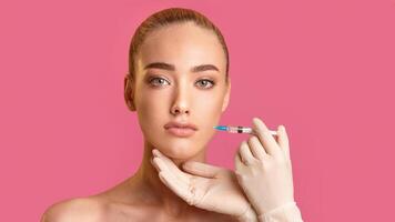 A woman with blonde hair is receiving a facial injection from a gloved hand. She is looking directly at the camera and has a neutral expression on her face. The background is a bright pink color. photo