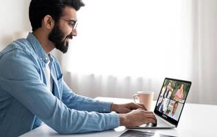 A young man is engaged in a virtual meeting using his laptop at his home office. He sits at a desk with a bright window in the background, interacting with multiple colleagues on the screen. photo