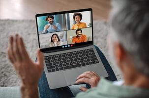Elderly man sits on a couch, using a laptop to participate in a group chat. The laptop screen shows four tiles of different individuals, each with a different background. photo
