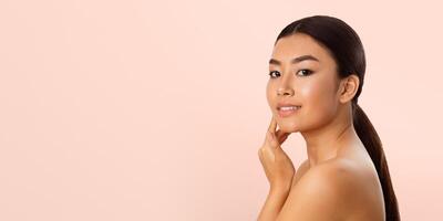 Asian young woman with long, black hair tied back in a ponytail. She is touching her face with one hand and looking directly at the camera with a soft smile. The background is a light pink color. photo