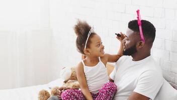 African American little girl, wearing a tiara, applies makeup to her fathers face with a brush while sitting on a bed. They are both smiling and having fun together photo