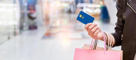 A woman standing in a shopping area, holding a credit card in one hand and multiple shopping bags in the other. She appears to be in the middle of a shopping spree, panorama with copy space photo