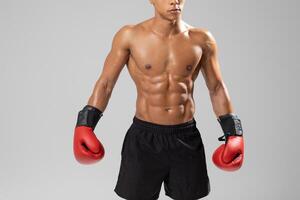 A fit young man boxer stands confidently in a studio, showcasing his defined abs and muscular arms while wearing vibrant red boxing gloves, cropped photo