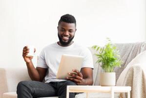 A happy African American man sits on a couch in a living room, holding a tablet in one hand and a mug of coffee in the other. He is smiling and looks relaxed. photo