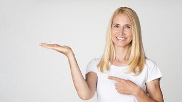 A happy blonde woman smiles and points to her open hand, presenting a product against a white background. Her expression is warm and inviting, suggesting she is excited about the product photo