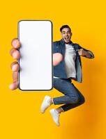 Handsome young Arab guy jumping on air, pointing at cellphone with empty screen on orange studio background. Millennial Muslim man demonstrating mockup for mobile app design photo