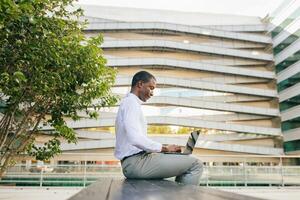 African American man in a white shirt and gray pants is sitting on a bench outside a modern office building. He is working on a laptop, looking at the screen with a focused expression photo