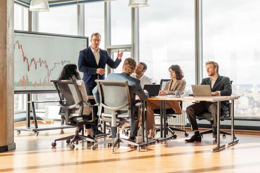 A group of business professionals are gathered around a conference table in a modern office. A presenter stands in front of the group, pointing to a large screen displaying a financial chart photo