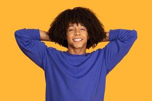 This image shows a young man with an curly hairstyle and a relaxed posture. He is smiling and leaning back with his hands behind his head. The background is a solid yellow color. photo