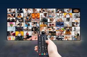 A hand holds a black remote control in front of a large screen displaying numerous images. The screen shows a grid of small videos or images depicting various scenes and activities photo