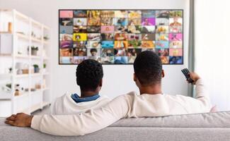 Two men are relaxing on a couch in a living room, watching television. The television is displaying a large, colorful grid of images. photo