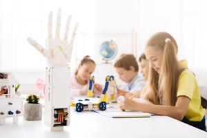 A robotic hand model stands in the foreground of the image, in focus, while a group of children work on a project in the background. The classroom is bright and filled with natural light. photo