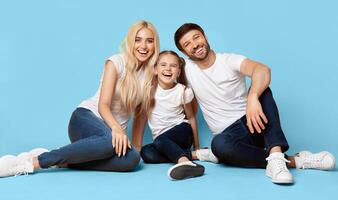 A happy family of three, a mother, father, and daughter, sit together on a light blue background. The family is wearing casual clothing and smiling broadly at the camera. photo