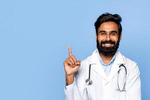 Indian man doctor, wearing a white lab coat with a stethoscope around his neck, smiles and points upward with his index finger. He has a dark beard and dark hair. The background is a solid blue color. photo