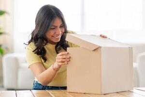 Excited middle eastern woman is standing at a table, opening a box that is placed in front of her. She appears to be focused on the contents of the box, her hands delicately handling the item inside. photo