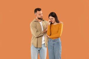 A man stands next to a distressed woman, offering her comfort and support. He places a reassuring hand on her shoulder while she wipes tears from her eyes. They are dressed in casual photo