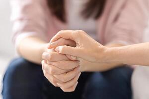 Two individuals sit closely, offering solace through hand-holding. Their intertwined hands signify support and empathy, creating a comforting atmosphere indoors, cropped photo