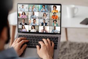A man is sitting on a couch and using a laptop to participate in a conference. The screen of the laptop is displaying a grid of feeds showing several different people photo