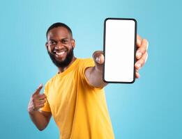 Excited black guy in casual wear pointing at cellphone with empty screen on blue studio background, space for website or mobile app design. Cellphone display mockup. Selective focus photo
