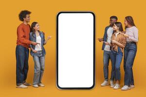 A group of five friends stand in front of a large smartphone with a blank white screen. They are all pointing at the screen, suggesting they are interested in what is being displayed photo